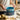 A person is lifting a blue non-stick round cake pan out of a stack, with a few pieces of food and a pot in the background, indicating it is used for baking.