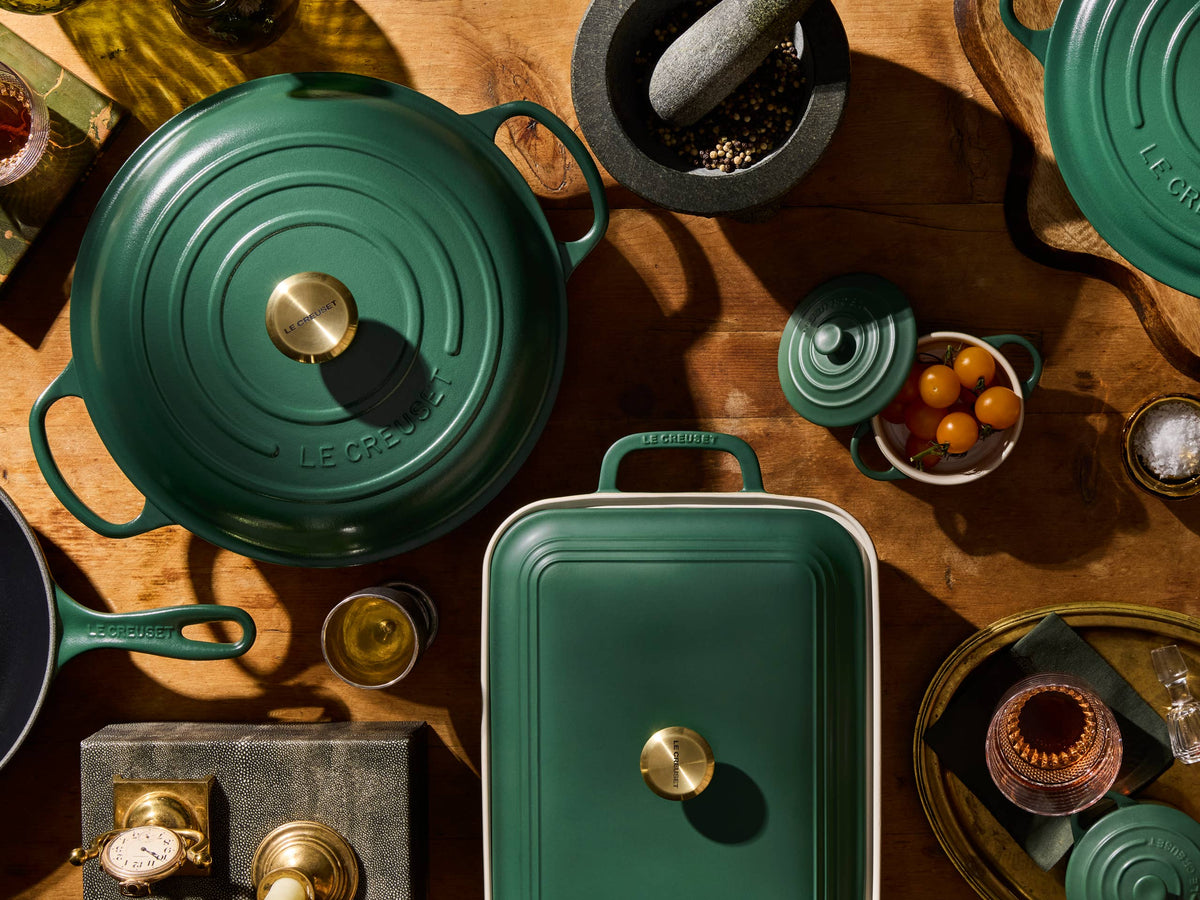 Green Le Creuset cookware on a wooden table, including a round lidded pot, a skillet and a baking dish