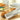 A perforated French bread pan made of aluminized steel, with a loaf of bread partially placed in it, set on a table with kitchen background.
